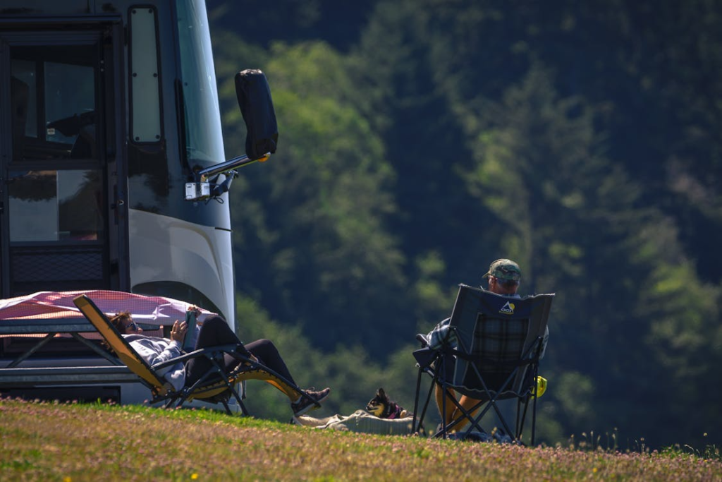Campers relaxing beside an RV in a peaceful campground surrounded by trees and natural scenery.